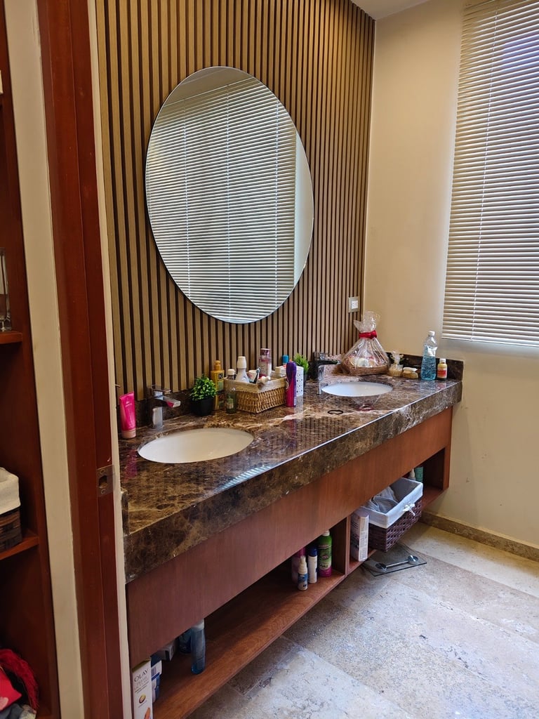 Modern bathroom vanity with dark granite countertop, round mirror, vertical striped wallpaper, and open shelving with various toiletries and supplies
